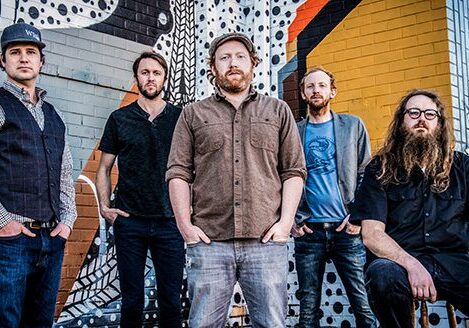 Five men posing against colorful graffiti wall.