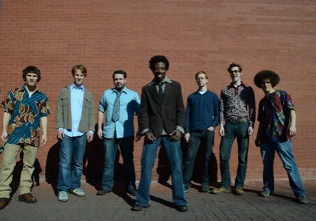 Seven men posing against a brick wall.