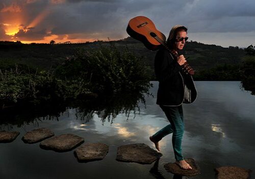 Person crossing river at sunset with guitar.