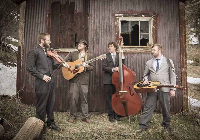 Four musicians playing instruments near rustic building.