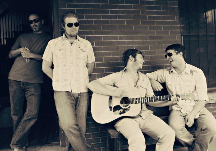 Four men with guitar, sitting and smiling.