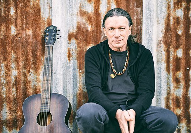 Man sitting with guitar against rustic wall.