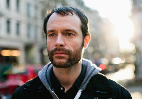 Man standing outdoors, city background, sunlight behind.