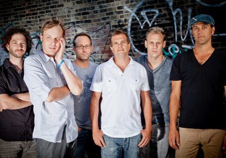 Six men standing against graffiti-covered wall.