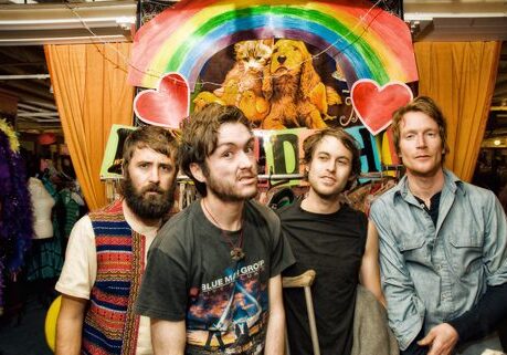 Four men standing under colorful rainbow banner.