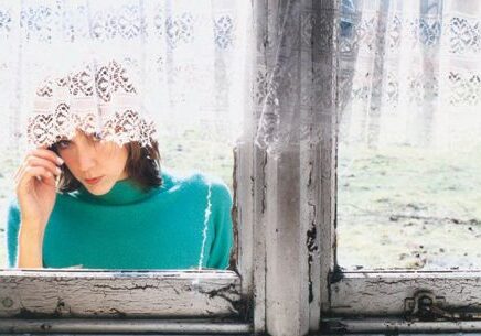 Woman peeking through lace curtain by window.