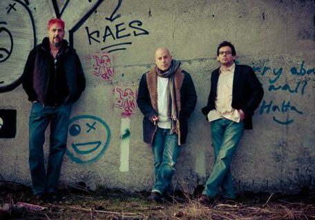 Three men standing against graffiti-covered wall.