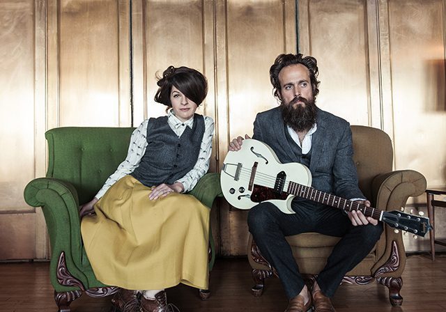 Man with guitar and woman sitting indoors.