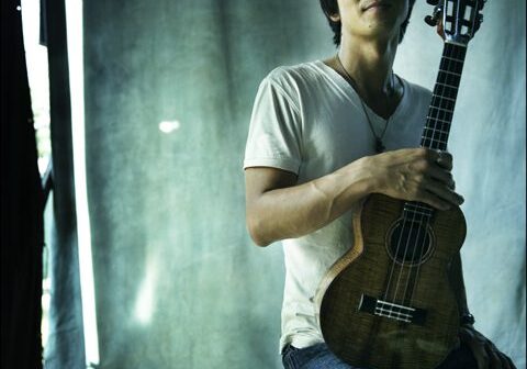 Person seated, holding a ukulele, smiling confidently.