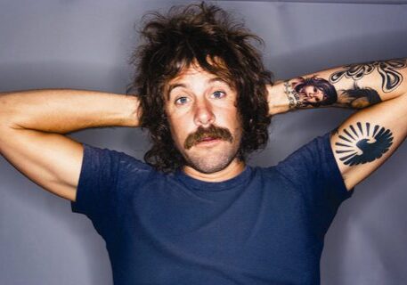 Man with tattoos and mustache, arms raised.