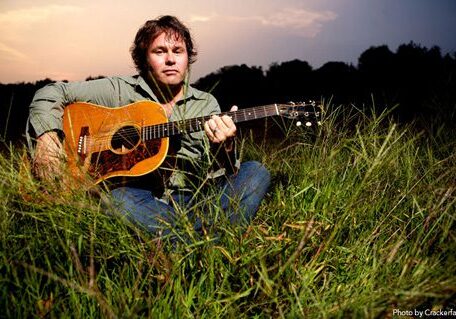 Man playing guitar in grassy field at sunset.