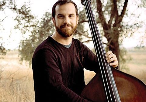 Man playing double bass outdoors near trees.