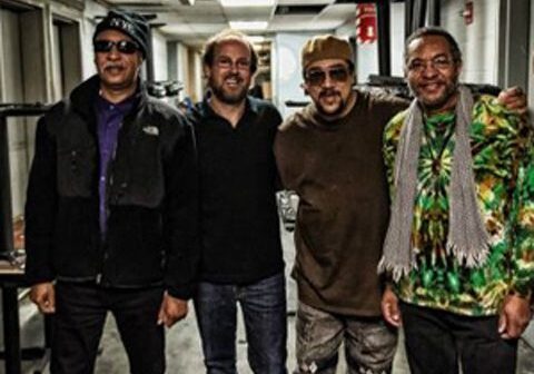 Four men standing together, smiling indoors.
