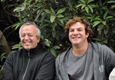 Two smiling men in front of greenery.