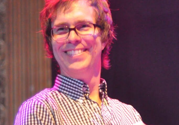 Person smiling on stage with glasses.