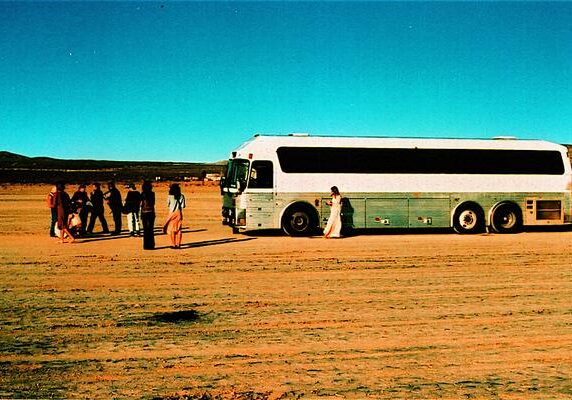 Bus on desert road with people nearby.