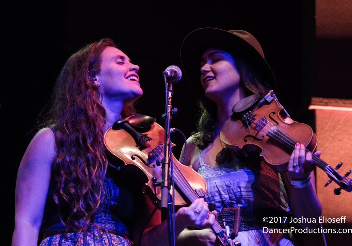 Two women joyfully playing violins on stage.