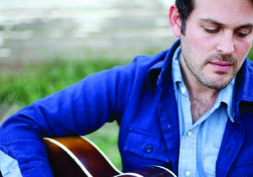 Man playing guitar outdoors, wearing blue jacket.