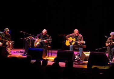 Band performing on stage with guitars, seated.