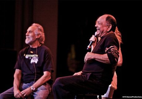 Two men laughing on stage with microphone.