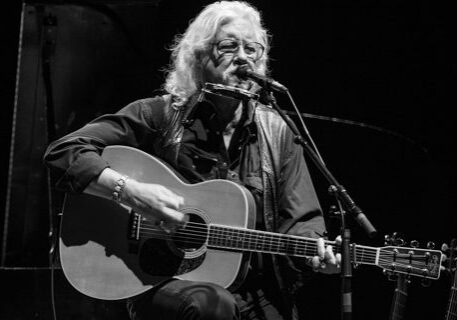 Musician playing guitar and harmonica on stage.