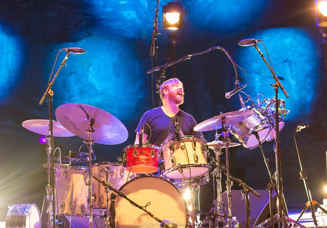 Drummer performing energetically on stage, blue lighting.