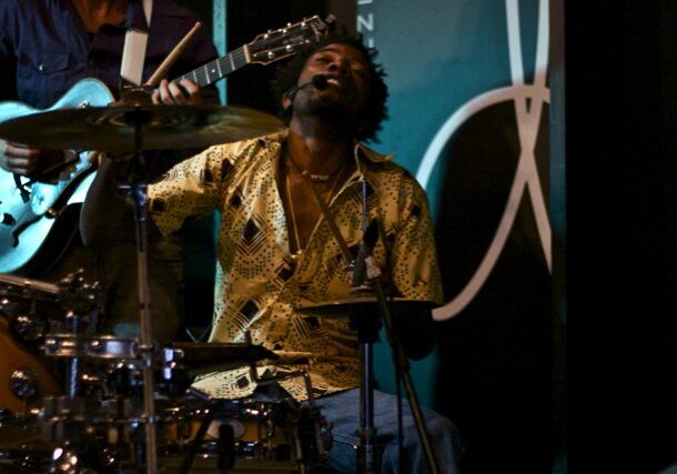 Musician passionately playing drums and guitar onstage.