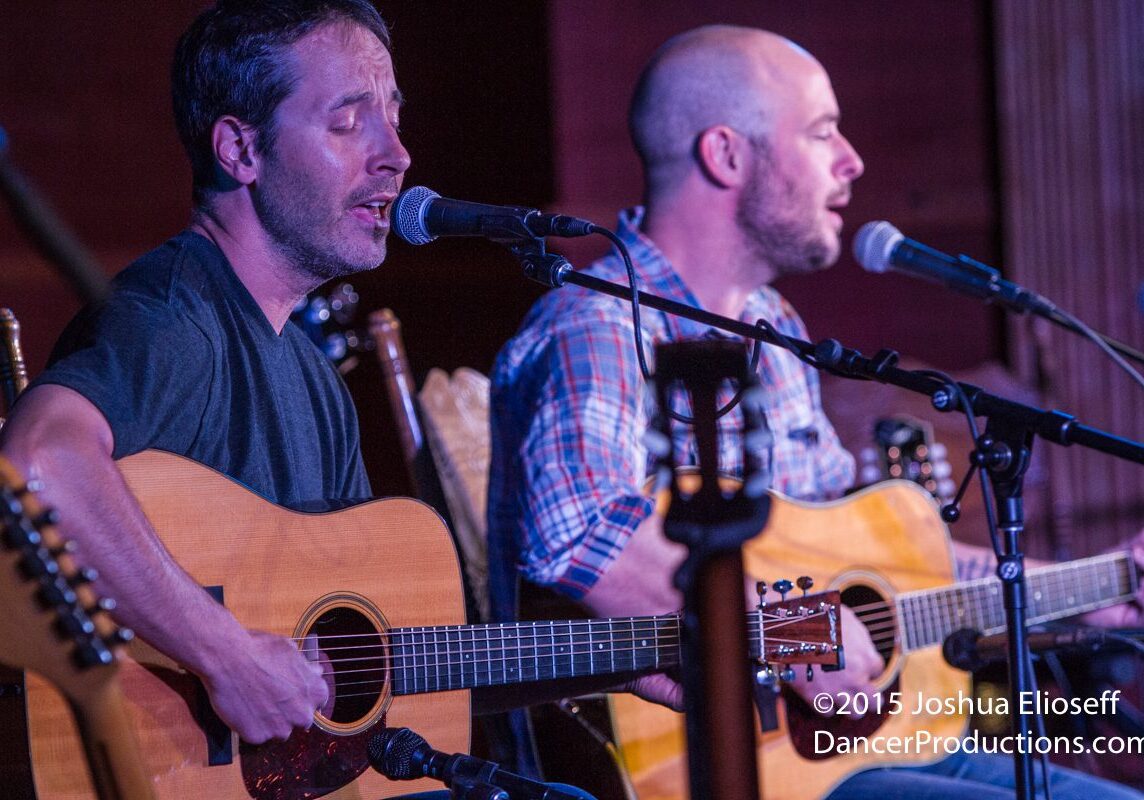 Two men performing with acoustic guitars onstage.