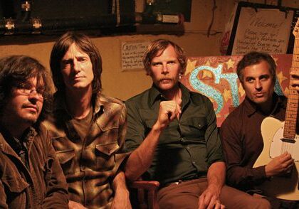 Four men sitting with a guitar indoors.