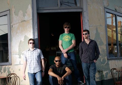 Four men posing outside a rustic building.