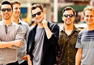 Five smiling men standing outdoors, wearing sunglasses.