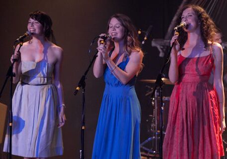 Three women singing on stage with microphones.
