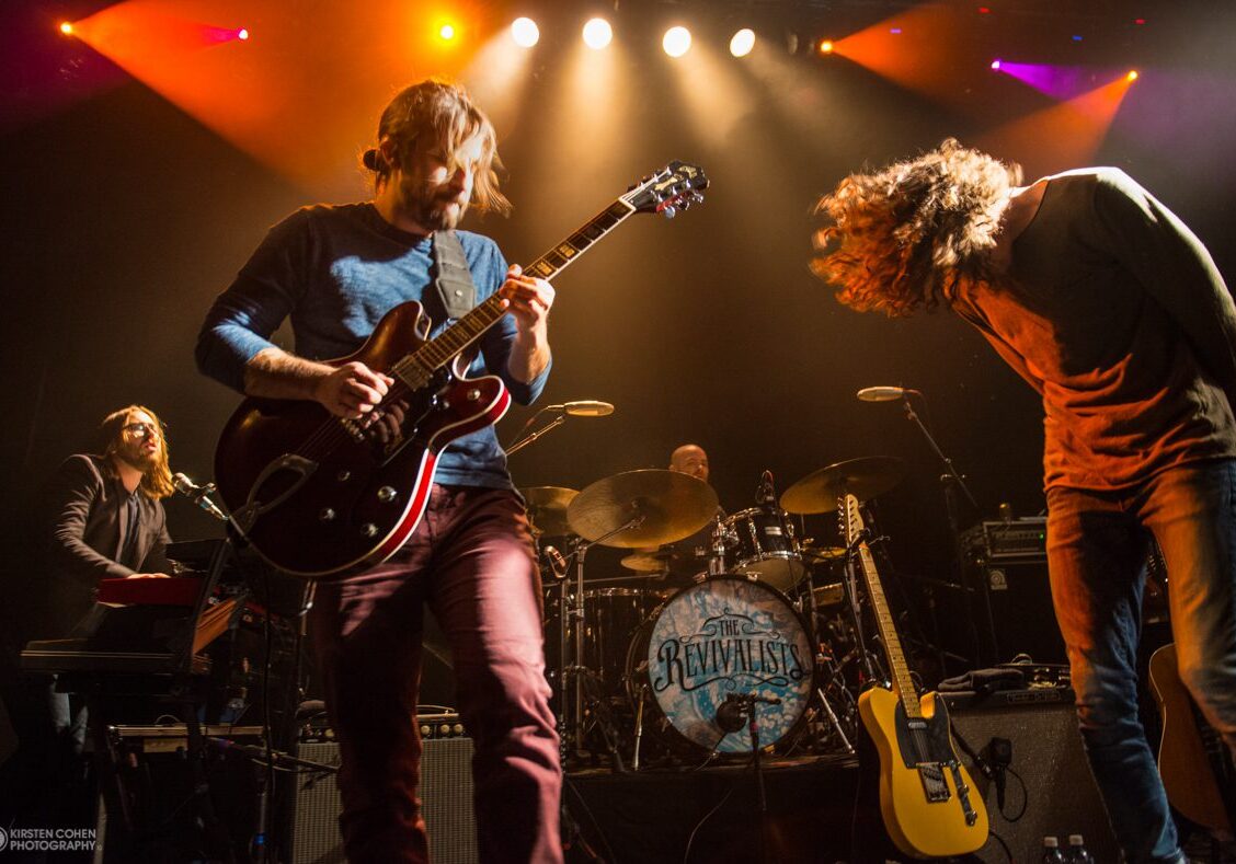 Band performing energetically on stage with lights.