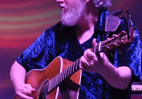 Guitarist performing on stage with colorful backdrop.