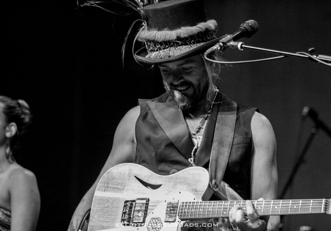Musician playing guitar on stage, wearing hat.