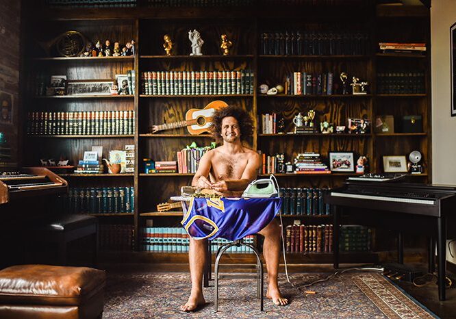 Shirtless man ironing in a library.