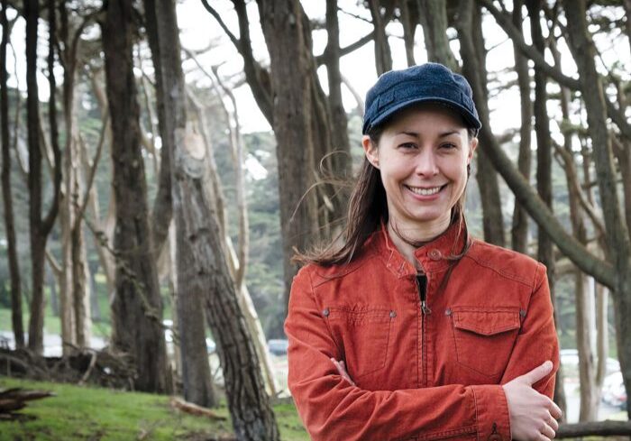 Woman smiling in forest, wearing orange jacket.
