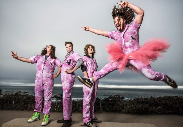 Band in pink outfits, one jumping energetically.