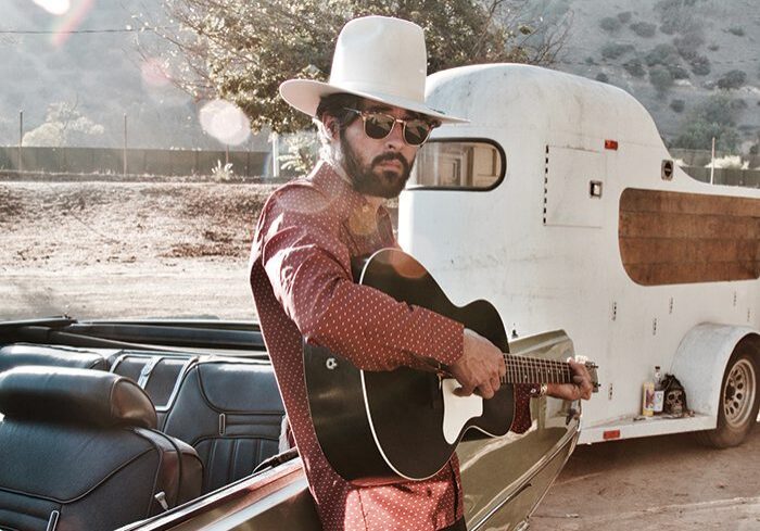 Man with guitar near vintage car and trailer.